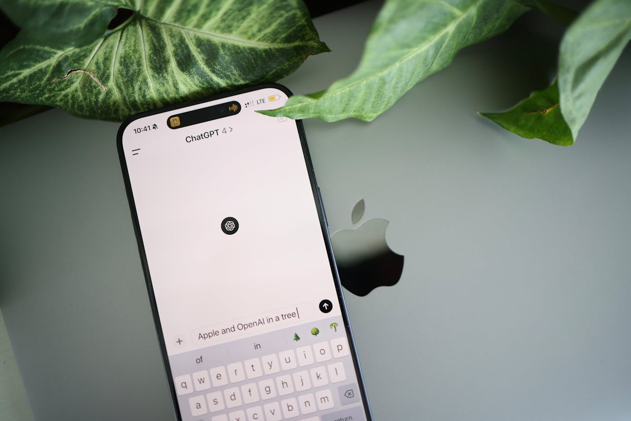 A smartphone shows a ChatGPT interface placed on an Apple laptop in a leafy environment.